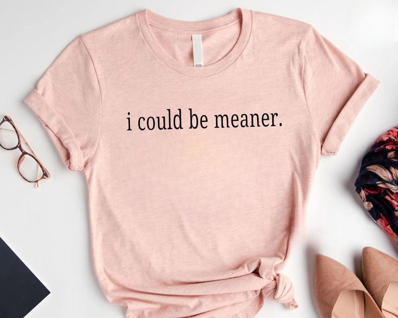 May include: A light pink t-shirt with the phrase "i could be meaner" printed in black. The shirt has short sleeves and a round neckline. The image also shows a pair of glasses, a scarf, and a pair of shoes.