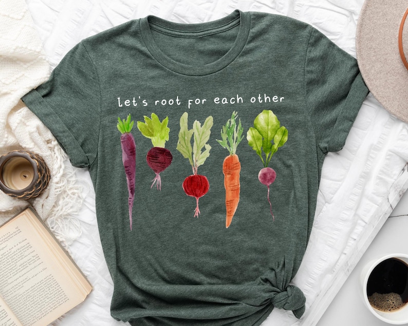 May include: Dark green t-shirt featuring a watercolor illustration of various root vegetables, including carrots and beets, with green leafy tops. The text "Let's root for each other" is printed above the vegetables. The shirt is knotted at the bottom.