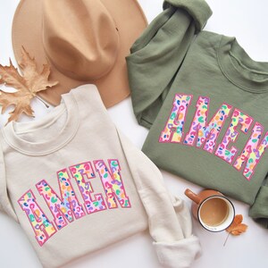Puede incluir: Dos sudaderas de color crema y verde oliva con la palabra "AMEN" en letras de estampado de leopardo rosa, arco&iacute;ris y azul. Las sudaderas est&aacute;n dispuestas sobre una superficie blanca con un sombrero marr&oacute;n, hojas de oto&ntilde;o y una taza de caf&eacute;.