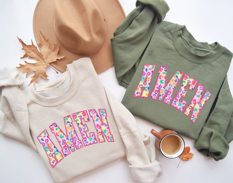 Puede incluir: Dos sudaderas de color crema y verde oliva con la palabra "AMEN" en letras de estampado de leopardo rosa, arco&iacute;ris y azul. Las sudaderas est&aacute;n dispuestas sobre una superficie blanca con un sombrero marr&oacute;n, hojas de oto&ntilde;o y una taza de caf&eacute;.