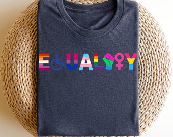 May include: Dark blue t-shirt with the word "EQUALITY" in colorful letters. Each letter is designed with symbols and colors representing different communities, including the LGBTQ+ community, disability rights, and more. The shirt is folded on a woven surface.
