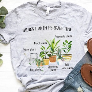 以下が含まれることがあります： 「Things I do in my spare time」というテキストと、「Propagate plants」、「Repot plants」、「Water plants」、「Buy plants」、「Rearrange plants」、「Talk with plants」などの植物関連の活動のリストが書かれたグレーのTシャツ。テキストは鉢植えの植物のイラストで囲まれています。