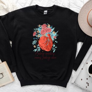 May include: Black sweatshirt featuring a graphic of a red heart surrounded by blue and green flowers. The text "Feeling emotions means feeling alive" is printed below the heart.