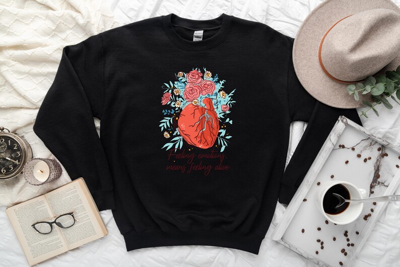 May include: Black sweatshirt featuring a graphic of a red heart surrounded by blue and green flowers. The text "Feeling emotions means feeling alive" is printed below the heart.