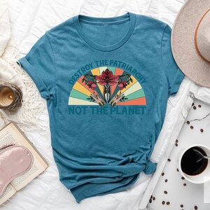 Op de afbeelding: Een tealblauw t-shirt met een retro-grafische print die "Vernietig het patriarchaat, niet de planeet" zegt. De grafiek toont een gestileerde baarmoeder met bloemen en een regenboog.
