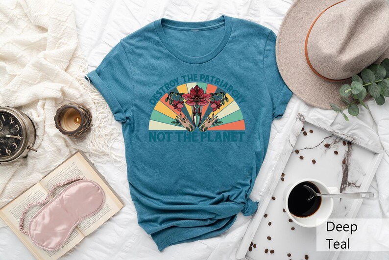 Op de afbeelding: Een tealblauw t-shirt met een retro-grafische print die "Vernietig het patriarchaat, niet de planeet" zegt. De grafiek toont een gestileerde baarmoeder met bloemen en een regenboog.
