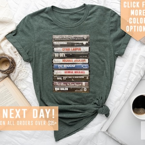 May include: A green t-shirt with a stack of vintage cassette tapes featuring artists like Madonna, Cyndi Lauper, Michael Jackson, Pat Benatar, George Michael, Van Halen, and more.