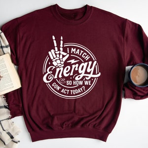 Op de afbeelding: Bordeauxkleurige trui met een witte afbeelding van een skelet hand die een vredesteken maakt en de tekst "I match energy & so how we gon' act today?"