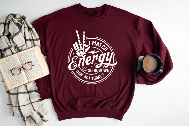 Op de afbeelding: Bordeauxkleurige trui met een witte afbeelding van een skelet hand die een vredesteken maakt en de tekst "I match energy & so how we gon' act today?"