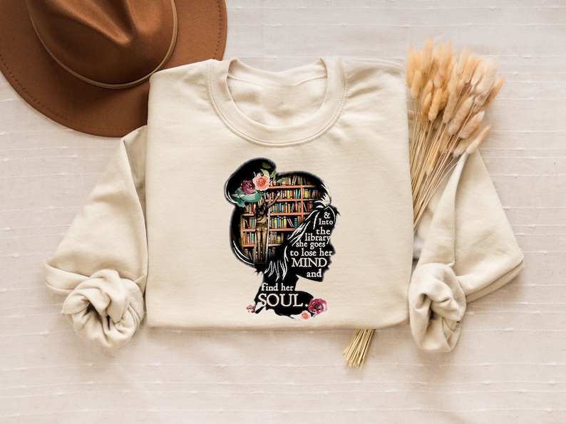 May include: A beige sweatshirt with a graphic of a woman's silhouette with a bookshelf for a head. The text reads "& Into the library she goes to lose her MIND and find her SOUL."