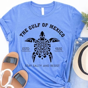 May include: Light blue t-shirt with a black graphic of a turtle and the text "THE GULF OF MEXICO ESTD 1550 STAY SALTY AND RESIST". The shirt is folded, with sandals and a hat visible.
