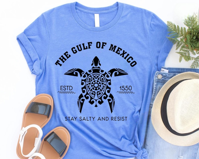 May include: Light blue t-shirt with a black graphic of a turtle and the text "THE GULF OF MEXICO ESTD 1550 STAY SALTY AND RESIST". The shirt is folded, with sandals and a hat visible.