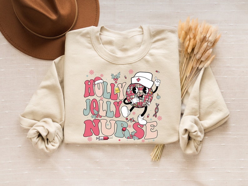 Op de afbeelding: Een cr&egrave;mekleurige sweatshirt met de woorden "HOLLY JOLLY NURSE" in een retro lettertype. Het ontwerp toont een discobal-personage met een verpleegstersmuts. Hulst- en sneeuwvlokaccenten maken het feestelijke ontwerp compleet.