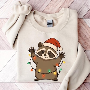 Puede incluir: Una sudadera color crema con un mapache de dibujos animados que lleva un gorro de Pap&aacute; Noel y est&aacute; envuelto en luces navide&ntilde;as de colores. El mapache tiene un cuerpo marr&oacute;n, una m&aacute;scara negra y sonr&iacute;e con los brazos levantados.