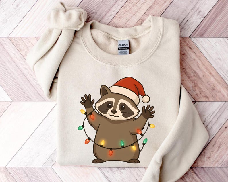 Puede incluir: Una sudadera color crema con un mapache de dibujos animados que lleva un gorro de Pap&aacute; Noel y est&aacute; envuelto en luces navide&ntilde;as de colores. El mapache tiene un cuerpo marr&oacute;n, una m&aacute;scara negra y sonr&iacute;e con los brazos levantados.