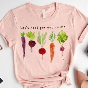 May include: A light pink t-shirt with the text "Let's root for each other" above a watercolor illustration of various root vegetables. The vegetables include a purple carrot, a beet, a radish, a carrot, and a turnip.