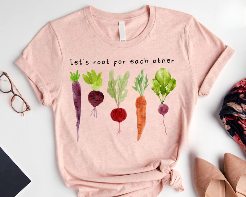 May include: A light pink t-shirt with the text "Let's root for each other" above a watercolor illustration of various root vegetables. The vegetables include a purple carrot, a beet, a radish, a carrot, and a turnip.