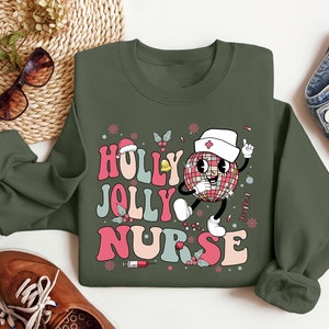 Op de afbeelding: Een olijfgroene sweatshirt met de woorden "Holly Jolly Nurse" in een retro lettertype. Het ontwerp toont een cartoon discobal karakter met een verpleegsterspet en een vinger omhoog. Feestelijke elementen zoals sneeuwvlokken en hulst zijn inbegrepen.