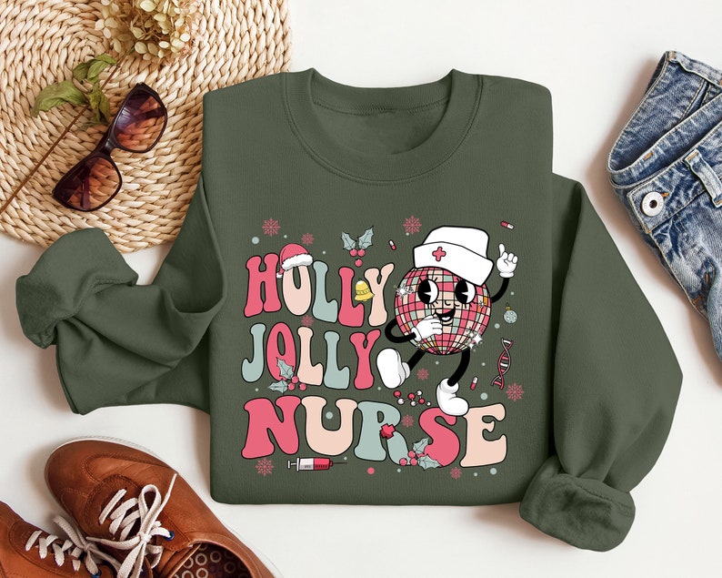 Op de afbeelding: Een olijfgroene sweatshirt met de woorden "Holly Jolly Nurse" in een retro lettertype. Het ontwerp toont een cartoon discobal karakter met een verpleegsterspet en een vinger omhoog. Feestelijke elementen zoals sneeuwvlokken en hulst zijn inbegrepen.