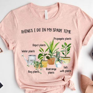 以下が含まれることがあります： 「THINGS I DO IN MY SPARE TIME」というテキストと、鉢植えの植物のグラフィックが描かれたピンクのTシャツ。植物の手入れに関するテキストが書かれており、「Repot plants」、「Propagate plants」、「Water plants」、「Buy plants」、「Rearrange plants」、「Talk with plants」などがあります。