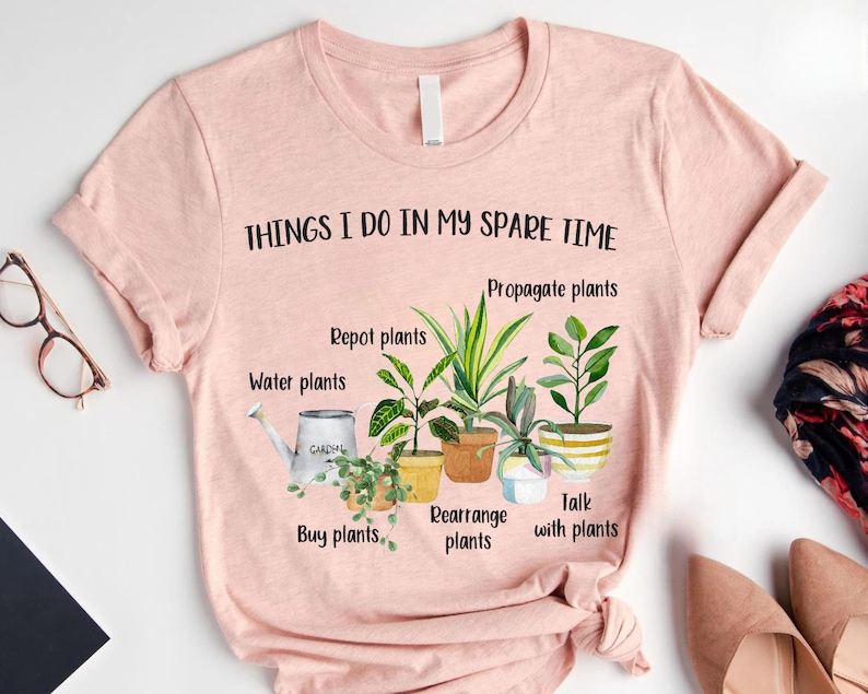 以下が含まれることがあります： 「THINGS I DO IN MY SPARE TIME」というテキストと、鉢植えの植物のグラフィックが描かれたピンクのTシャツ。植物の手入れに関するテキストが書かれており、「Repot plants」、「Propagate plants」、「Water plants」、「Buy plants」、「Rearrange plants」、「Talk with plants」などがあります。