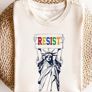 May include: A white t-shirt with a graphic of the Statue of Liberty holding a sign that says "RESIST" in rainbow colors.