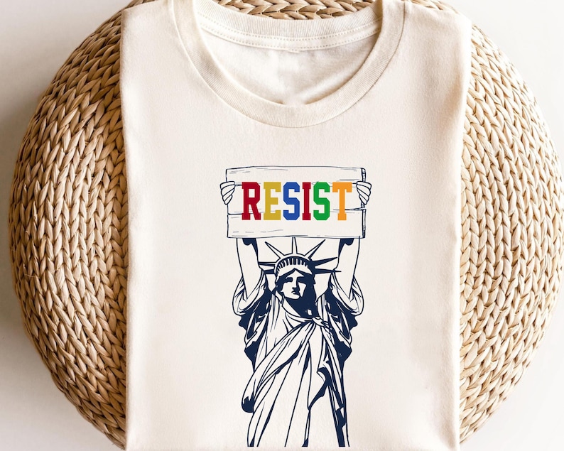May include: A white t-shirt with a graphic of the Statue of Liberty holding a sign that says "RESIST" in rainbow colors.