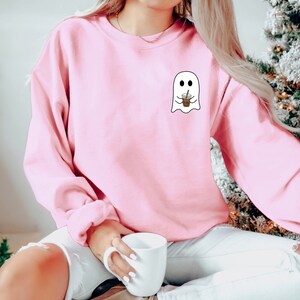 Puede incluir: Sudadera rosa claro con un peque&ntilde;o gr&aacute;fico de un fantasma sosteniendo una taza de caf&eacute;. El fantasma tiene dos ojos negros y una boca sonriente. La sudadera es de color liso y tiene mangas largas.