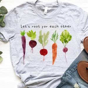 May include: A light gray t-shirt featuring a watercolor design of various root vegetables, including carrots and beets. The text "Let's root for each other" is printed above the vegetables. The shirt is paired with blue jeans and brown sandals.