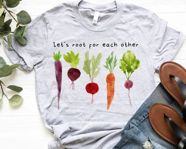 May include: A light gray t-shirt featuring a watercolor design of various root vegetables, including carrots and beets. The text "Let's root for each other" is printed above the vegetables. The shirt is paired with blue jeans and brown sandals.