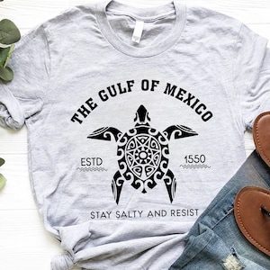 May include: A light gray t-shirt featuring a black graphic of a turtle with the text "THE GULF OF MEXICO" above it. Below the turtle are the words "ESTD 1550" and "STAY SALTY AND RESIST". Brown sandals and blue jeans are visible.