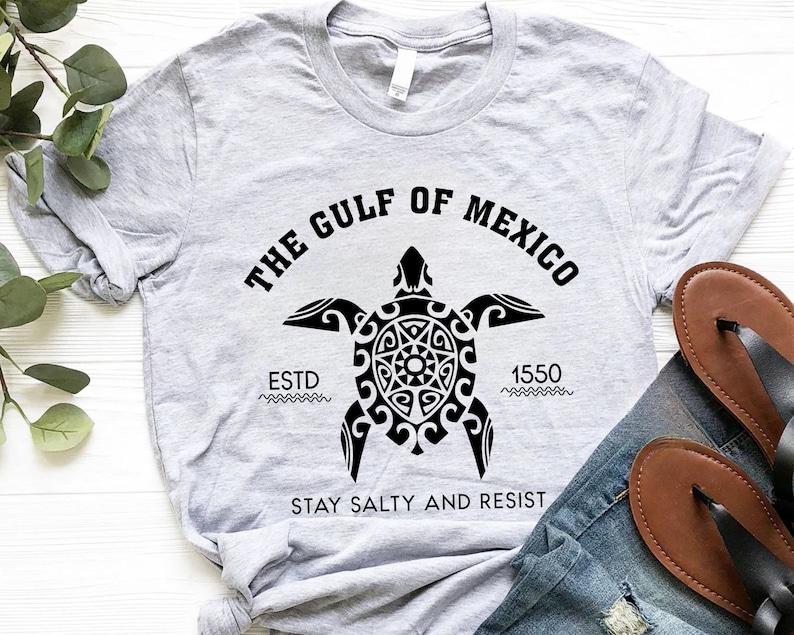 May include: A light gray t-shirt featuring a black graphic of a turtle with the text "THE GULF OF MEXICO" above it. Below the turtle are the words "ESTD 1550" and "STAY SALTY AND RESIST". Brown sandals and blue jeans are visible.