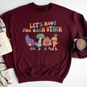 May include: A maroon sweatshirt with the text "Let's root for each other" in a colorful retro font. The text is surrounded by cartoon images of a carrot, eggplant, broccoli, and tomato.