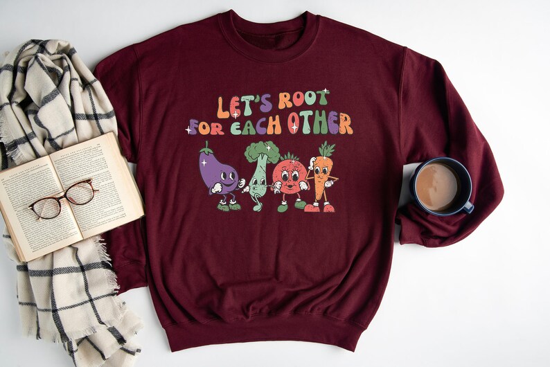 May include: A maroon sweatshirt with the text "Let's root for each other" in a colorful retro font. The text is surrounded by cartoon images of a carrot, eggplant, broccoli, and tomato.