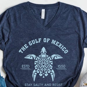 May include: Navy blue V-neck t-shirt featuring a light blue graphic of a sea turtle with the text "THE GULF OF MEXICO" and "ESTD 1550". The shirt also includes the phrase "STAY SALTY AND RESIST".