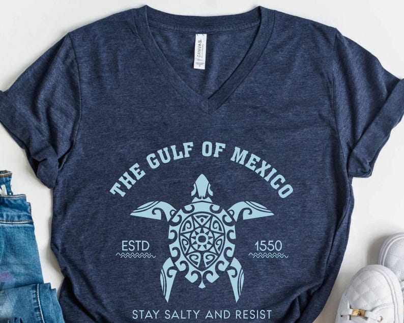 May include: Navy blue V-neck t-shirt featuring a light blue graphic of a sea turtle with the text "THE GULF OF MEXICO" and "ESTD 1550". The shirt also includes the phrase "STAY SALTY AND RESIST".
