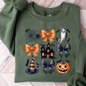 May include: Green sweatshirt featuring a Halloween design. The design includes orange bows, a haunted house, a ghost, a witch's hat, a spider web, bats, and a jack-o'-lantern. The sweatshirt is folded on a wooden surface.