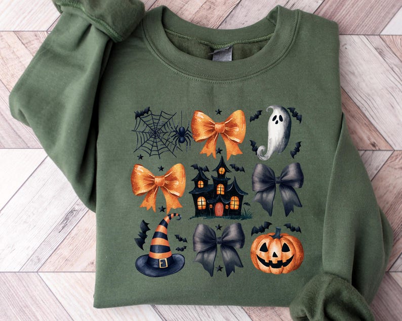May include: Green sweatshirt featuring a Halloween design. The design includes orange bows, a haunted house, a ghost, a witch's hat, a spider web, bats, and a jack-o'-lantern. The sweatshirt is folded on a wooden surface.