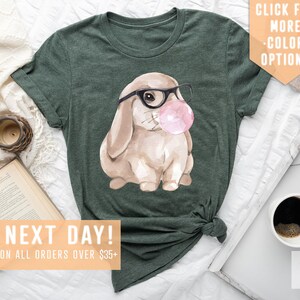 May include: A green t-shirt with a watercolor illustration of a brown bunny wearing glasses and blowing a pink bubble. The text "SHIPS NEXT DAY! FREE SHIPPING ON ALL ORDERS OVER $35+" is at the bottom of the image.