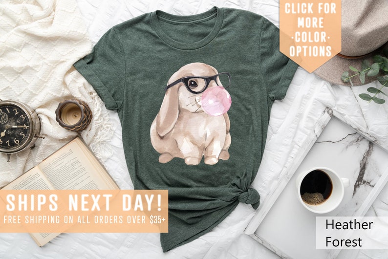 May include: A green t-shirt with a watercolor illustration of a brown bunny wearing glasses and blowing a pink bubble. The text "SHIPS NEXT DAY! FREE SHIPPING ON ALL ORDERS OVER $35+" is at the bottom of the image.