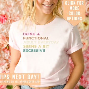 May include: A pink t-shirt with the text "BEING A FUNCTIONAL ADULT EVERYDAY SEEMS A BIT EXCESSIVE" in a rainbow gradient. The text is centered on the shirt. The shirt is being worn by a person. The text "SHIPS NEXT DAY! FREE SHIPPING ON ALL ORDERS OVER $35+" is at the bottom of the image. The text "CLICK FOR MORE -COLOR- OPTIONS" is in the top right corner of the image.