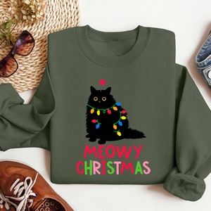May include: A green sweatshirt with a black cat wearing Christmas lights and the text "MEOWY CHRISTMAS" in red, green, and pink.