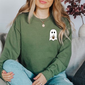 Puede incluir: Sudadera verde oliva con un dise&ntilde;o de fantasma blanco sosteniendo una taza de caf&eacute;. El fantasma tiene ojos negros y una boca sonriente. La sudadera de manga larga est&aacute; hecha de un material suave. El dise&ntilde;o est&aacute; en el pecho izquierdo.