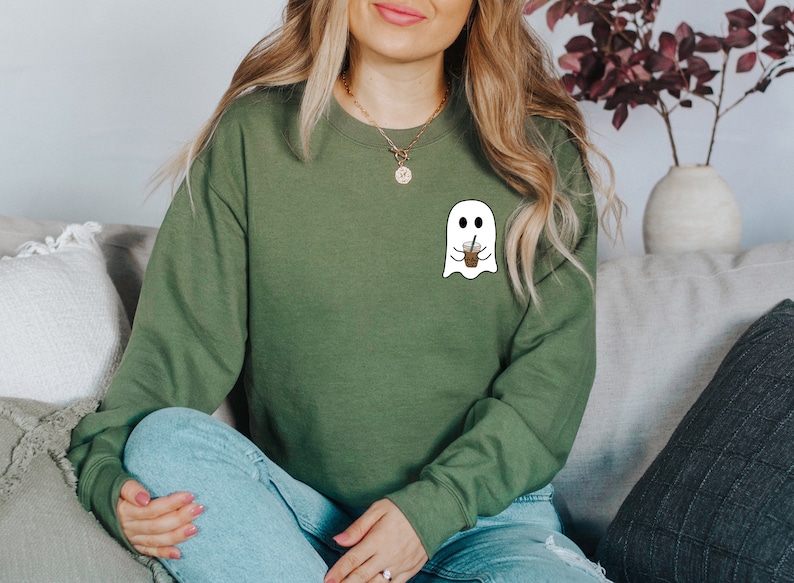 Puede incluir: Sudadera verde oliva con un dise&ntilde;o de fantasma blanco sosteniendo una taza de caf&eacute;. El fantasma tiene ojos negros y una boca sonriente. La sudadera de manga larga est&aacute; hecha de un material suave. El dise&ntilde;o est&aacute; en el pecho izquierdo.