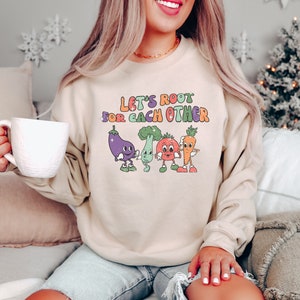 May include: A beige sweatshirt with the text "Let's Root For Each Other" in colorful letters. The text is surrounded by cartoon vegetables with faces.