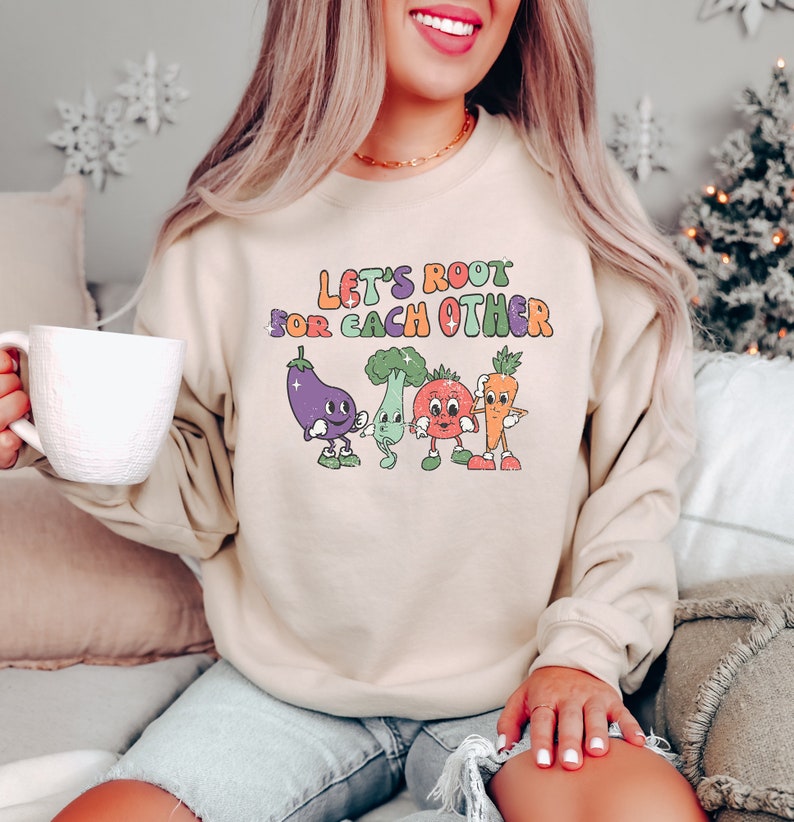 May include: A beige sweatshirt with the text "Let's Root For Each Other" in colorful letters. The text is surrounded by cartoon vegetables with faces.