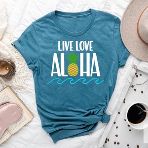 May include: Teal t-shirt with the words "LIVE LOVE ALOHA" in white, a yellow pineapple, and blue waves. The shirt is laid flat on a white surface, with a hat and coffee cup nearby. The shirt is a deep teal color.
