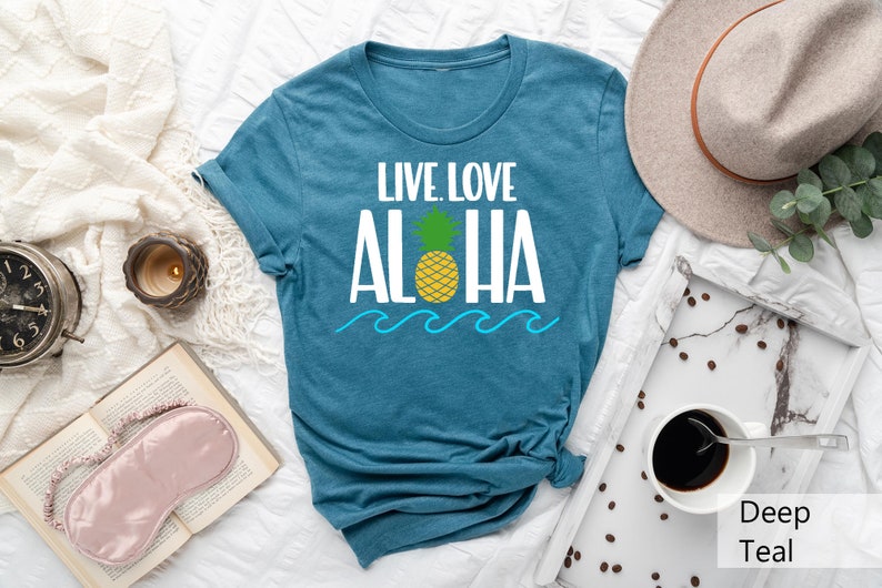 May include: Teal t-shirt with the words "LIVE LOVE ALOHA" in white, a yellow pineapple, and blue waves. The shirt is laid flat on a white surface, with a hat and coffee cup nearby. The shirt is a deep teal color.