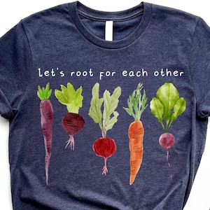 May include: A navy blue t-shirt with the text "Let's root for each other" above a watercolor illustration of various root vegetables. The vegetables include carrots, beets, and radishes.