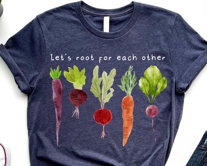 May include: A navy blue t-shirt with the text "Let's root for each other" above a watercolor illustration of various root vegetables. The vegetables include carrots, beets, and radishes.